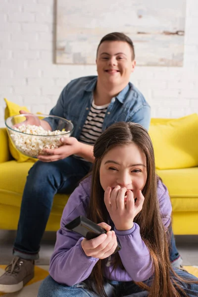Positive girl with down syndrome holding remote controller near blurred friend with popcorn at home — Stock Photo
