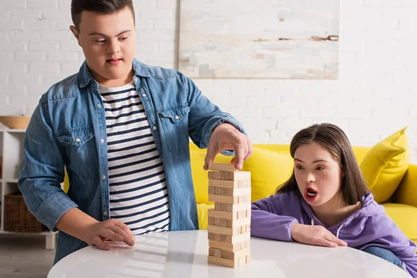 Niño con síndrome de Down jugando bloques de madera juego cerca de amigo sorprendido en casa - foto de stock