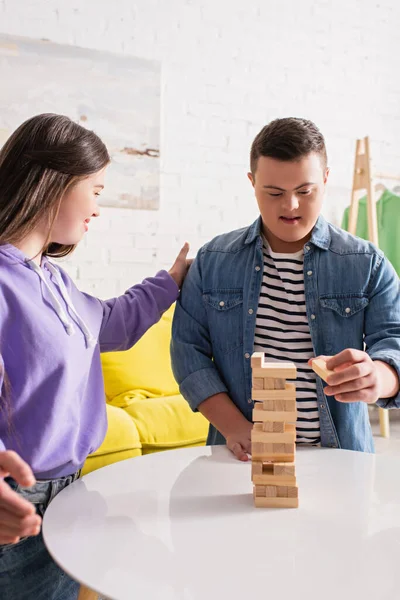 Menina sorridente com síndrome de down olhando amigo jogar blocos de madeira jogo em casa — Fotografia de Stock