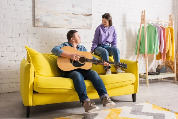 Adolescente tocando la guitarra acústica y mirando a un amigo con síndrome de Down en casa - foto de stock