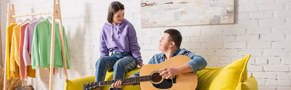 Дівчина-підліток з синдромом Дауна, що сидить поруч з другом, грає на акустичній гітарі вдома, банер — стокове фото