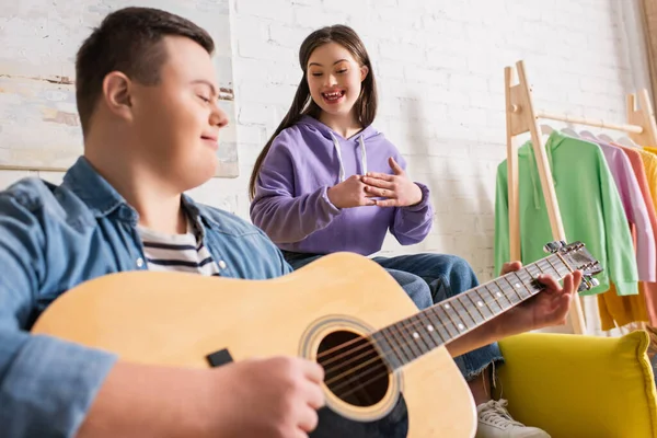 Веселая девушка с синдромом Дауна сидит рядом с размытым другом, играя на акустической гитаре дома — стоковое фото