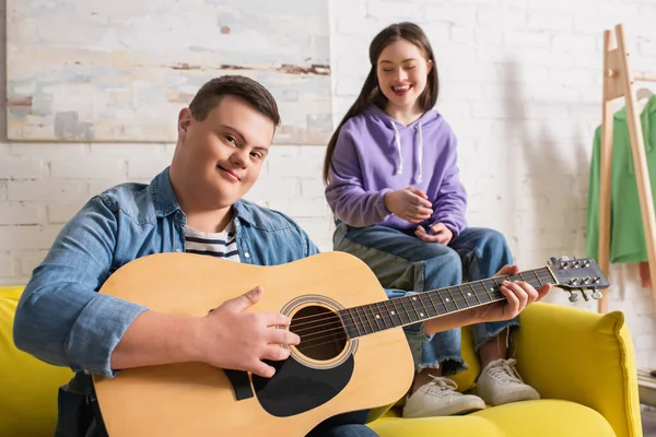 Мальчик с синдромом Дауна держит акустическую гитару и смотрит в камеру рядом с другом дома — стоковое фото