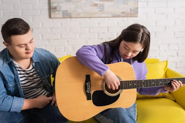 Chica con síndrome de Down tocando la guitarra acústica cerca de amigo en el sofá en casa - foto de stock