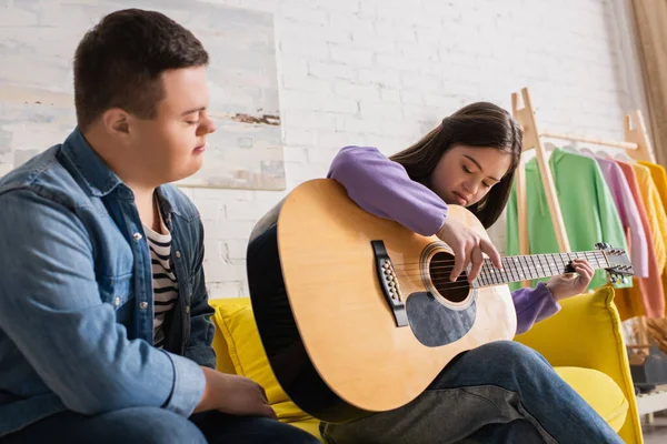 Adolescente ragazza con sindrome di Down suonare la chitarra acustica vicino amico sul divano — Foto stock