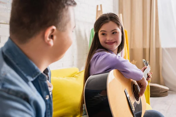 Smiling teenager with down syndrome playing acoustic guitar near blurred friend at home — Stock Photo