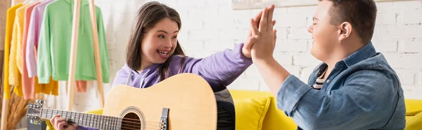 Cheerful teenagers with down syndrome and acoustic guitar giving high five at home, banner — Stock Photo