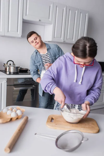 Teenager with down syndrome holding milk near friend cooking in kitchen — Stock Photo
