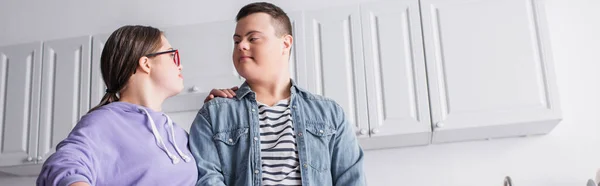 Low angle view of teenager with down syndrome looking at boyfriend in kitchen, banner — Stock Photo