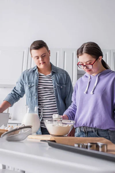 Chica adolescente con síndrome de Down sosteniendo la harina cerca de los ingredientes y amigo en la cocina - foto de stock