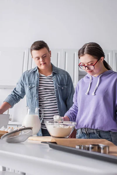 Menina adolescente com síndrome de down segurando farinha perto de ingredientes e amigo na cozinha — Fotografia de Stock