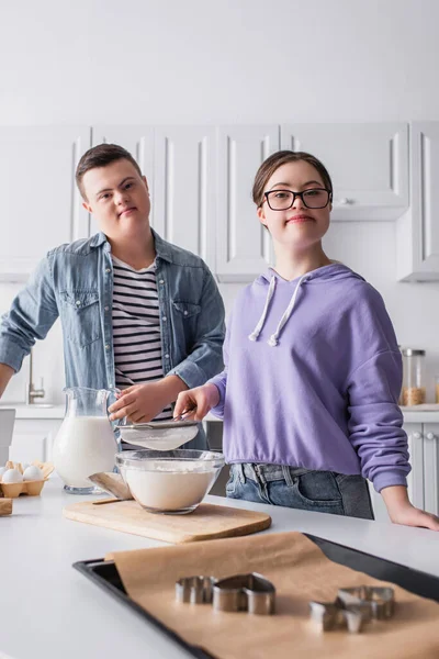 Adolescentes en síndrome de Down mirando la cámara cerca de la comida en la cocina - foto de stock