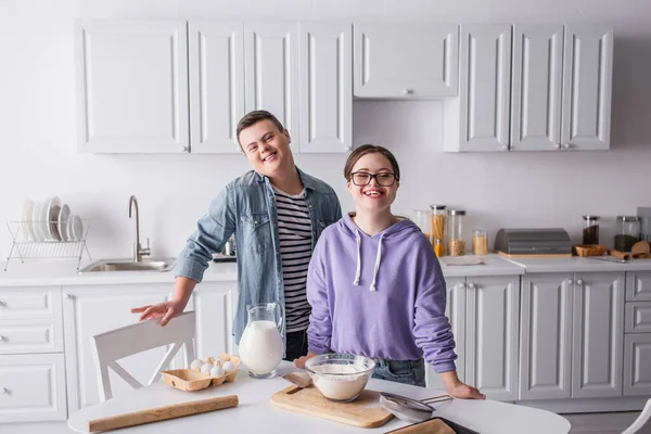 Adolescentes felizes com síndrome de down olhando para a câmera perto de comida na cozinha — Fotografia de Stock