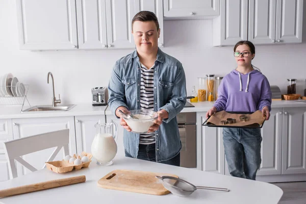 Adolescente com síndrome de down segurando farinha perto da namorada com assadeira na cozinha — Fotografia de Stock