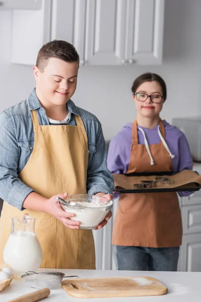 Adolescente sorridente com síndrome de down segurando farinha perto de namorada borrada com assadeira na cozinha — Fotografia de Stock