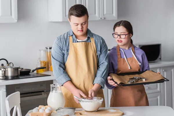 Teen couple with down syndrome in aprons cooking in kitchen — Stock Photo