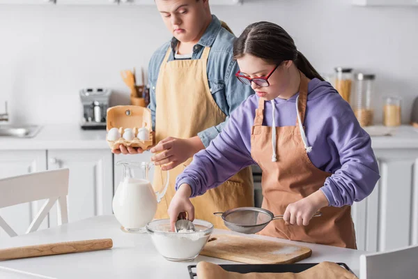 Menina com síndrome de down cozinhar perto de amigo segurando ovos na cozinha — Fotografia de Stock