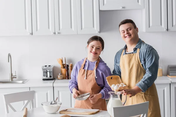 Positive teenagers with down syndrome in aprons cooking and looking at camera in kitchen — Stock Photo