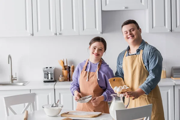 Adolescents positifs atteints du syndrome du duvet dans les tabliers de cuisine et regardant la caméra dans la cuisine — Photo de stock