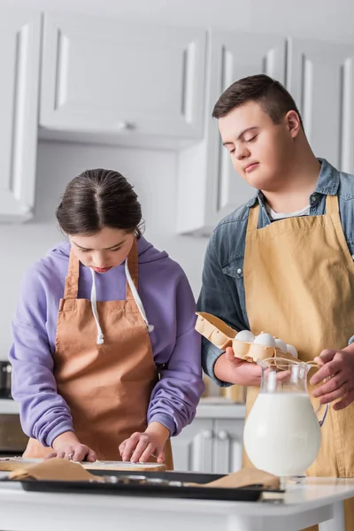 Chica con síndrome de Down cocinar cerca de amigo con huevos y bandeja para hornear en la cocina - foto de stock