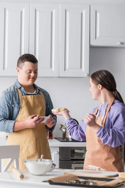 Adolescente positiva con síndrome de Down sosteniendo teléfono inteligente cerca de amigo con masa en la cocina - foto de stock