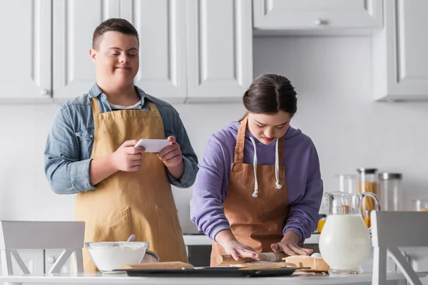 Adolescente com síndrome de down fazendo massa perto de ingredientes e amigo segurando smartphone na cozinha — Fotografia de Stock