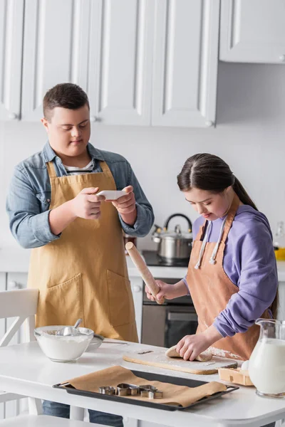 Teenager with down syndrome cooking dough near friend with smartphone at home — Stock Photo