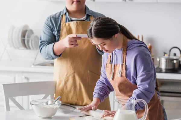 Teenager with down syndrome rolling dough near food and friend with smartphone — Stock Photo