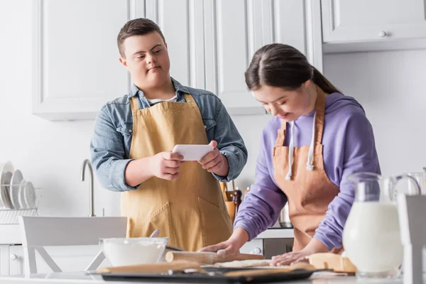 Adolescente con síndrome de Down sosteniendo teléfono inteligente mientras amigo borroso rodando masa en la cocina - foto de stock