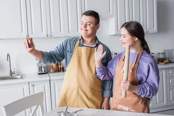 Улыбающиеся подростки с синдромом Дауна, имеющие видеозвонок на смартфоне во время приготовления пищи на кухне — стоковое фото