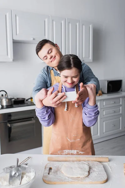 Adolescente com síndrome de down segurando celular perto de amigo em avental e massa na cozinha — Fotografia de Stock
