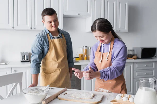 Smiling girl with down syndrome making dough near friend in apron in kitchen — Stock Photo