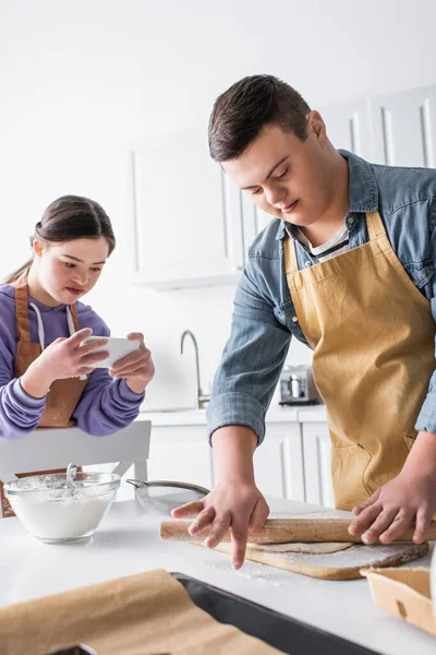 Teen boy with down syndrome rolling dough near friend with smartphone in kitchen — Stock Photo