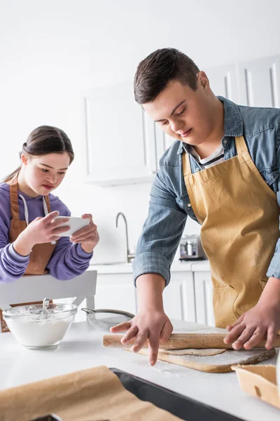 Adolescent garçon avec vers le bas syndrome roulant pâte près ami avec smartphone dans cuisine — Photo de stock