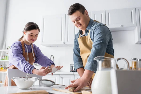 Smiling teenager with down syndrome rolling dough near friend taking photo in kitchen — Stock Photo