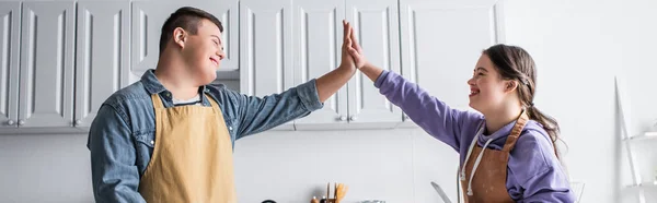 Cheerful friends with down syndrome in aprons giving high five in kitchen, banner — Stock Photo