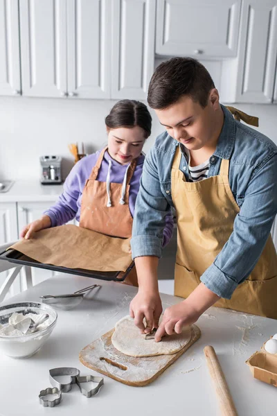 Adolescente com síndrome de down fazendo biscoito perto de amigo com assadeira na cozinha — Fotografia de Stock