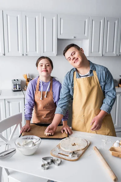 Amigos positivos com síndrome de down fazendo biscoito e olhando para a câmera na cozinha — Fotografia de Stock
