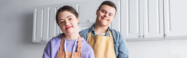 Positive couple with down syndrome in aprons looking at camera in kitchen, banner — Stock Photo