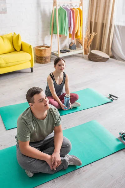 High angle view of teen boy with down syndrome sitting on fitness mat near friend at home — Stock Photo