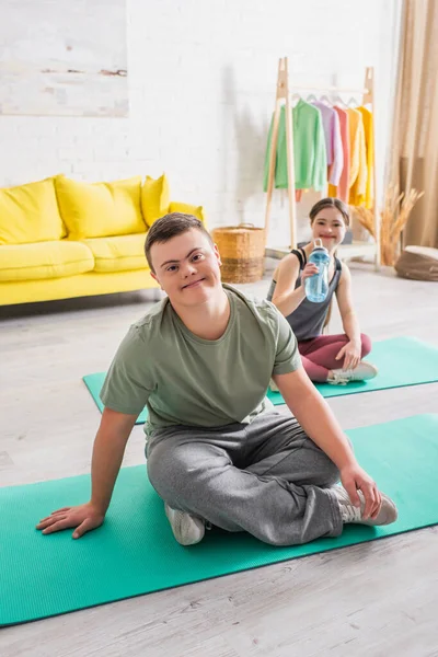 Adolescente sorridente com síndrome de down olhando para a câmera no tapete de fitness em casa — Fotografia de Stock