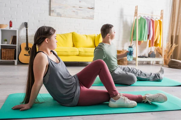 Side view of teenager with down syndrome sitting on fitness mat near friend at home — Stock Photo