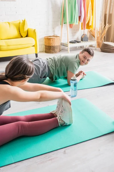 Adolescente positiva con síndrome de Down haciendo ejercicio en la alfombra de fitness cerca de un amigo en casa — Stock Photo