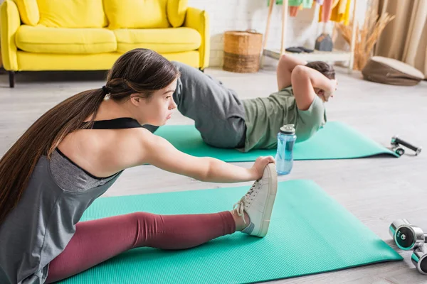 Side view of teenager with down syndrome training on fitness mat near dumbbells and friend at home — Stock Photo