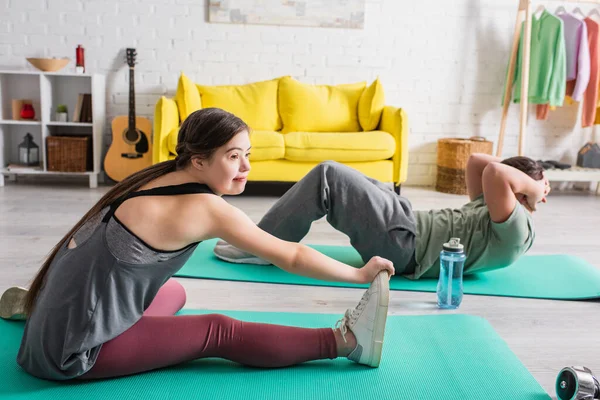 Teen girl with down syndrome training on fitness mat near blurred friend at home — Stock Photo