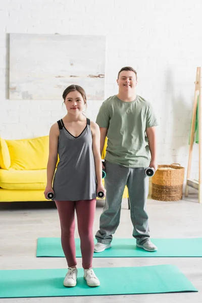 Adolescentes amigos com síndrome de down segurando halteres em tapetes de fitness em casa — Fotografia de Stock