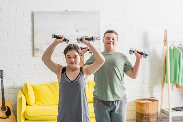 Teenager with down syndrome working out with dumbbells near blurred friend at home — Stock Photo