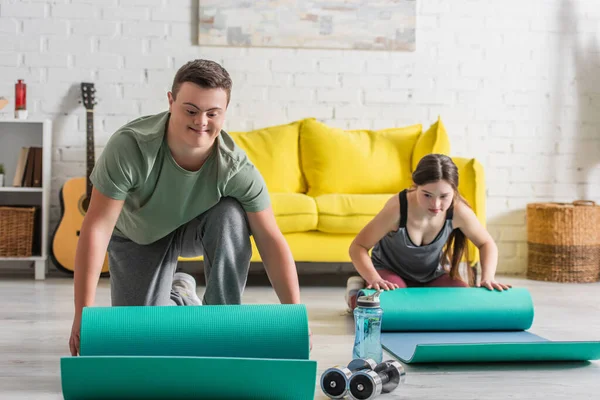 Sorrindo adolescente menino com síndrome de down segurando tapete de fitness perto de halteres e amigo em casa — Fotografia de Stock