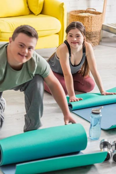 Adolescente con síndrome de Down sentado cerca de la esterilla de fitness y amigo borroso en casa - foto de stock