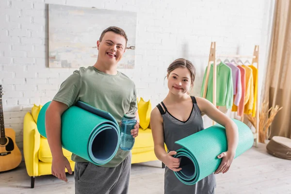 Adolescentes alegres con síndrome de Down sosteniendo tapetes de fitness y botella deportiva en casa - foto de stock